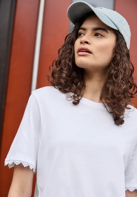 T-Shirt mit Crochet-Details an den Ärmeln White Frau mit lockigen Haaren trägt ein weißes T-Shirt und eine weiße Baseballkappe vor roter Wand.