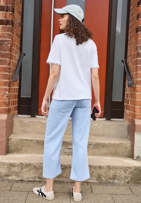 T-Shirt mit Crochet-Details an den Ärmeln White Frau von hinten in weißem T-Shirt, hellen Jeans und Mütze steht vor einer Backsteintür auf Stufen.