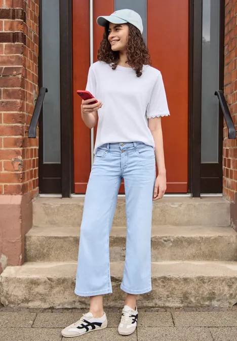 T-Shirt mit Crochet-Details an den Ärmeln White Frau in weißem T-Shirt, weiter Jeans und Sneakern mit rotem Smartphone auf Treppe vor roter Tür.