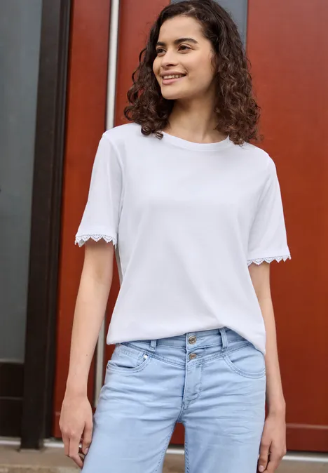 T-Shirt mit Crochet-Details an den Ärmeln White Frau in weißem T-Shirt und hellblauer Jeans vor rotem Hintergrund.