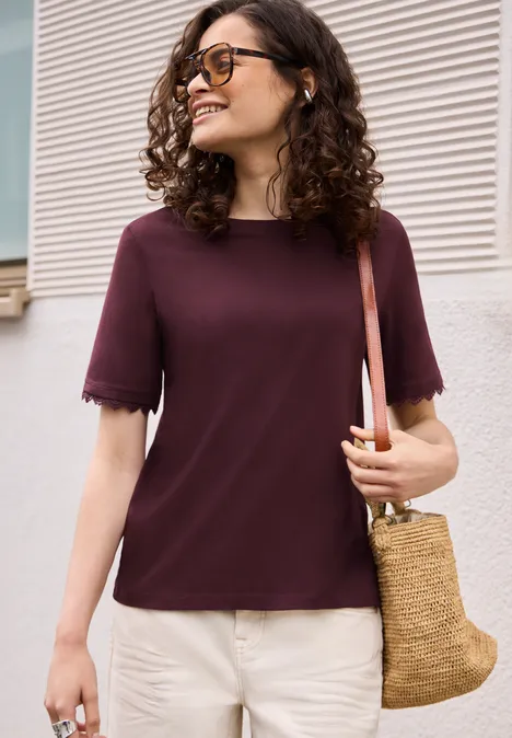 Frau mit hüftlanger, lockiger Frisur trägt bordeauxfarbenes T-Shirt, helle Hose und Rattantasche.