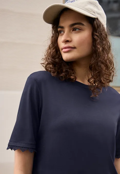 Persoon met krullend haar en pet draagt een donkerblauw T-shirt. Gezicht vaag.