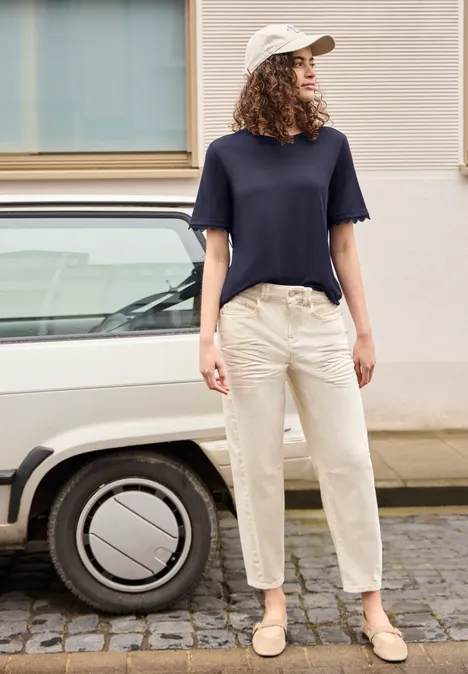 Vrouw met pet, blauw shirt en lichte broek staat naast witte auto op het trottoir.