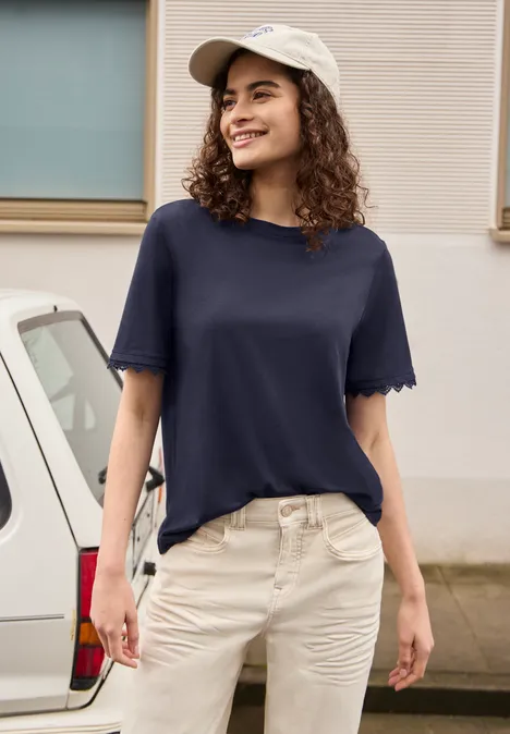 Een vrouw staat voor een auto, draagt een blauw T-shirt, een lichte broek en een witte pet.