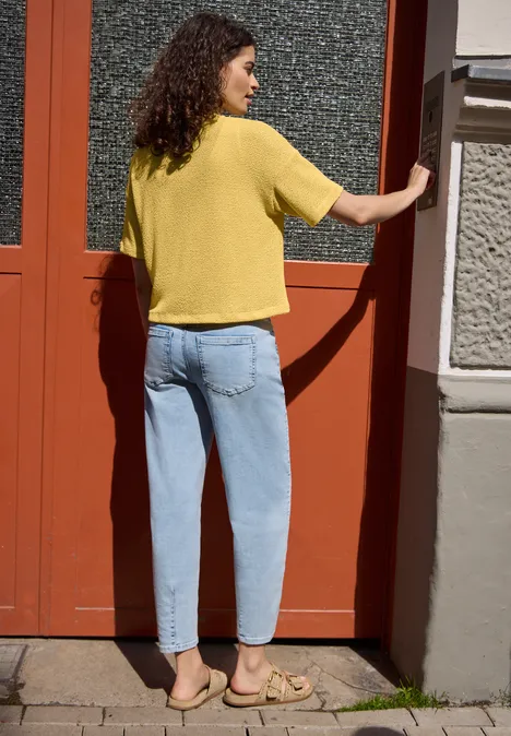 Eine Frau mit gelbem T-Shirt, blauen Jeans und Sandalen steht vor einer roten Tür und blickt zur Seite.
