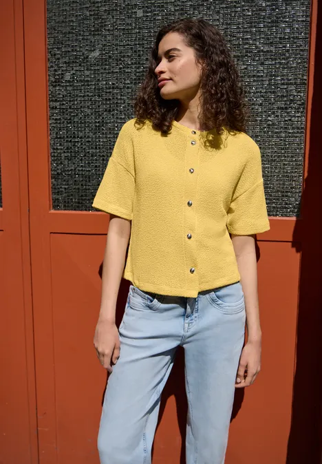 Eine Frau in gelbem Kurzarmshirt und blauen Jeans steht vor einer roten Tür.