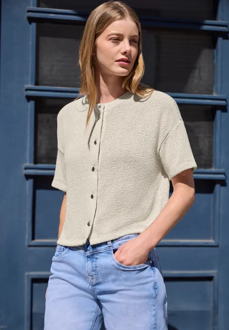 Eine Frau trägt eine kurzärmelige, helle Bluse und Jeans und steht vor einer blauen Tür.