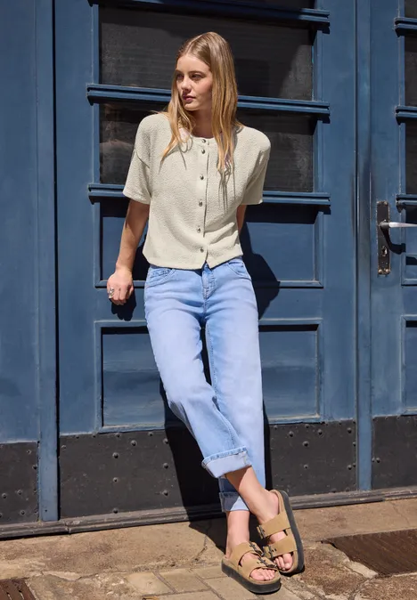 Kurzarm Jacke im Strick-Look mit Rundhals vanilla sand Frau lehnt vor einer blauen Tür, trägt helle Bluse, blaue Jeans und Sandalen.