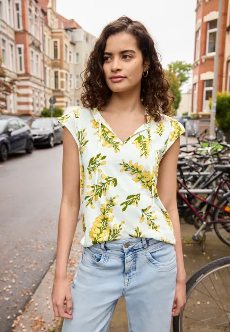Vrouw met krullend haar, draagt een blouse met citroenenprint en spijkerbroek, staand in een fietsenstraat.