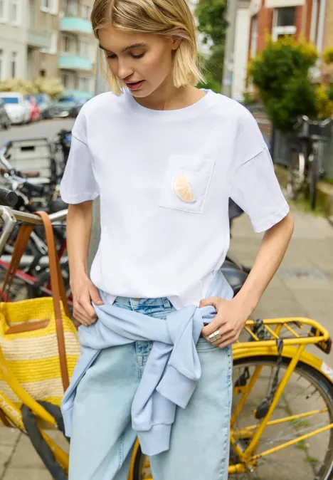 T-Shirt mit Rundhals und Perlendetail White Person trägt weißen T-Shirt und helle Jeans, mit hellblauer Jacke um die Taille und steht vor einem gelben Fahrrad.