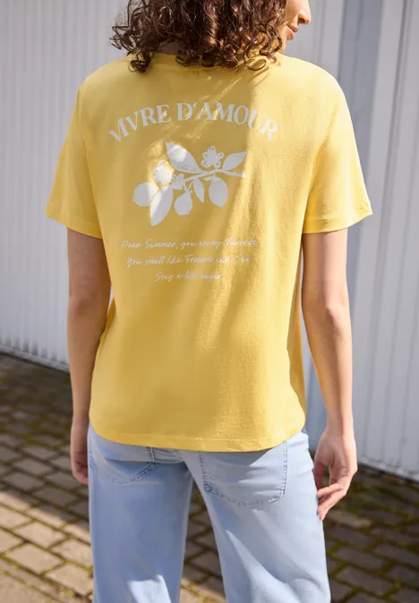 Eine Person trägt ein gelbes T-Shirt mit dem Aufdruck ‚VIVRE D'AMOUR‘ und einem weißen Blumenmotiv.
