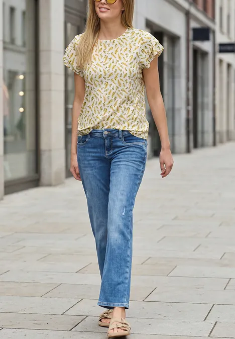 Eine Frau in gelbem Muster-Oberteil, blauem Jeans und Sandalen spaziert lächelnd in einer Einkaufsstraße.