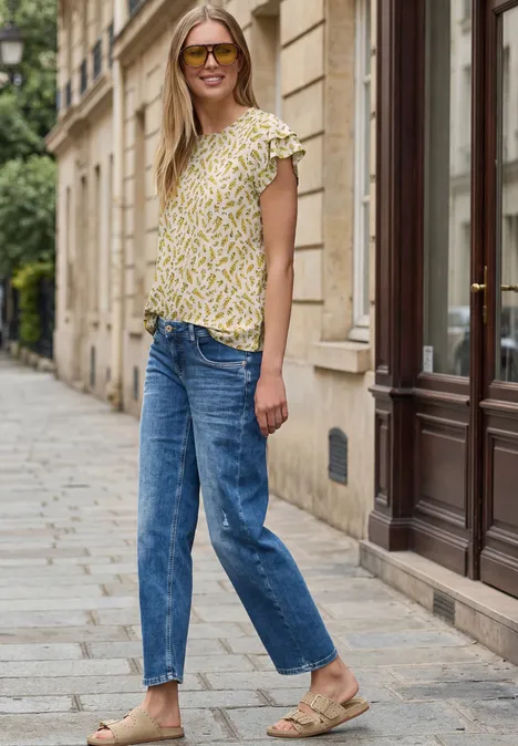 Frau in gelbem, gemustertem Top, blauen Jeans und Sandalen steht auf einem Gehweg vor Altbauten.