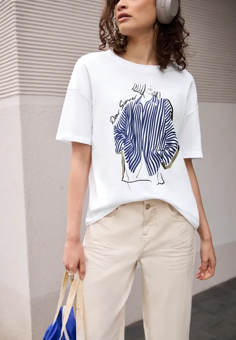 Persoon in beige broek en wit T-shirt met blauw lijnenschets-print, draagt koptelefoon en rieten tas.