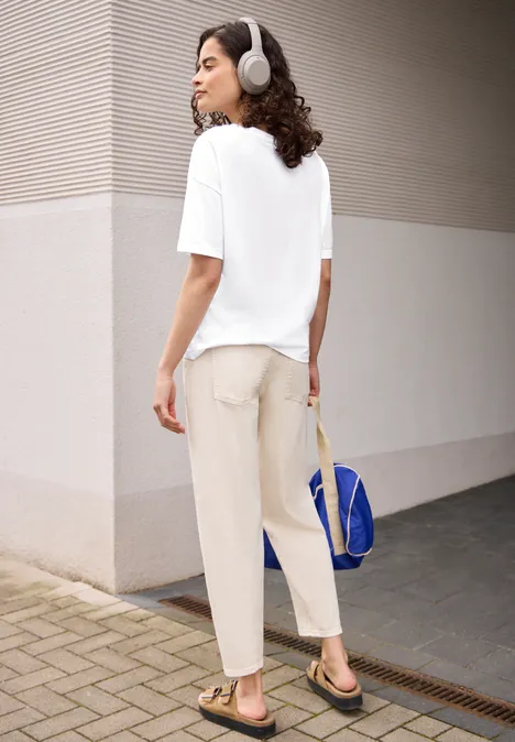 Persoon staat bij een gebouwhoek, draagt een wit t-shirt, beige broek, sandalen, koptelefoon en blauwe tas.