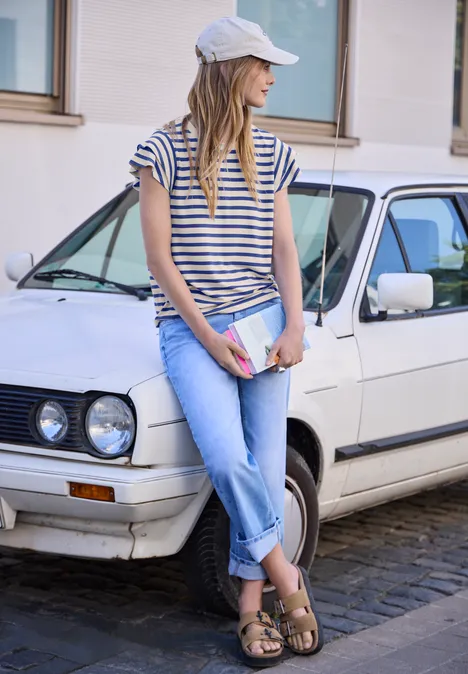 T-Shirt mit Volant-Ärmeln und Print optic blue Frau in Streifenshirt und Jeans lehnt an weißem Auto, trägt Cap und hält ein Buch.