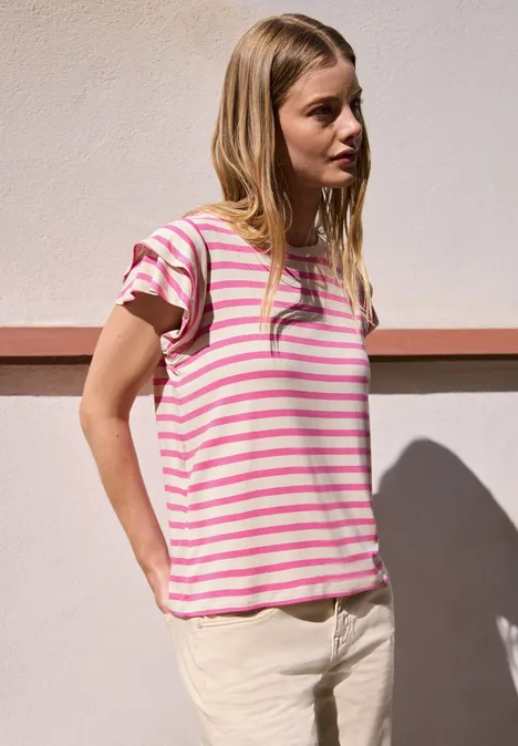 T-Shirt mit Volant-Ärmeln und Print simply pink Eine junge Frau steht vor einer hellen Wand, trägt pink-weiß gestreiftes T-Shirt und helle Hose.