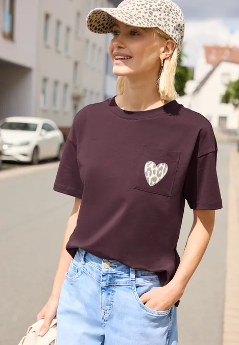 Person trägt dunkelrotes T-Shirt mit Herzmotiv, Jeans und Baseballcap, steht in ruhiger Wohnstraße.
