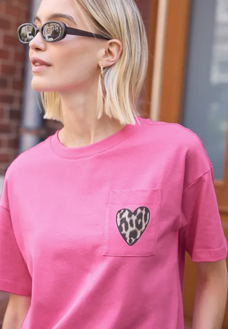 Frau mit kurzen blonden Haaren und Sonnenbrille trägt ein pinkes T-Shirt mit Herz-Leopardenmuster auf der Tasche.