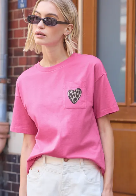 Person trägt pinkes T-Shirt mit Herz-Design auf Brusttasche und helle Jeans, stehend vor Backsteinwand.