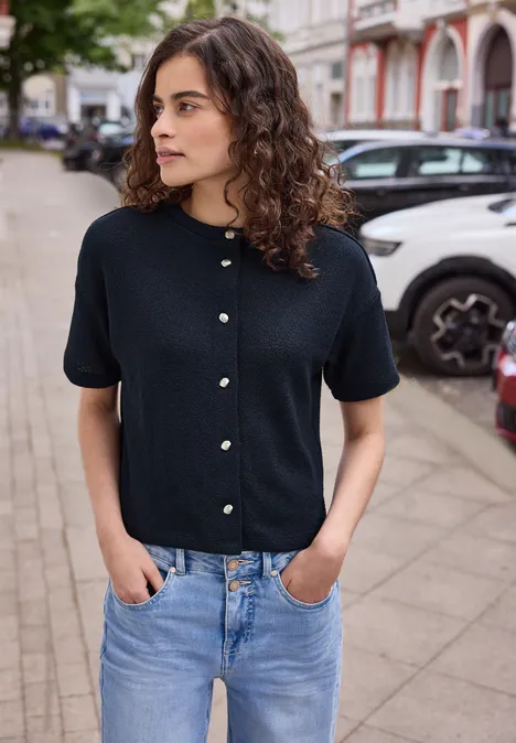 Kurzarm Jacke im Strick-Look mit Rundhals maritime blue Frau in schwarzem Kurzarmpullover mit Perlenknöpfen und hellblauen Jeans steht auf dem Gehweg vor parkenden Autos.