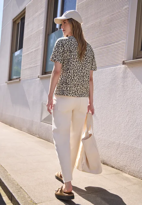 T-Shirt mit Rundhals und Leo-Muster light buffed beige Frau in gemustertem Shirt, hellen Hosen und Cap mit Tasche spaziert an weißer Hauswand.