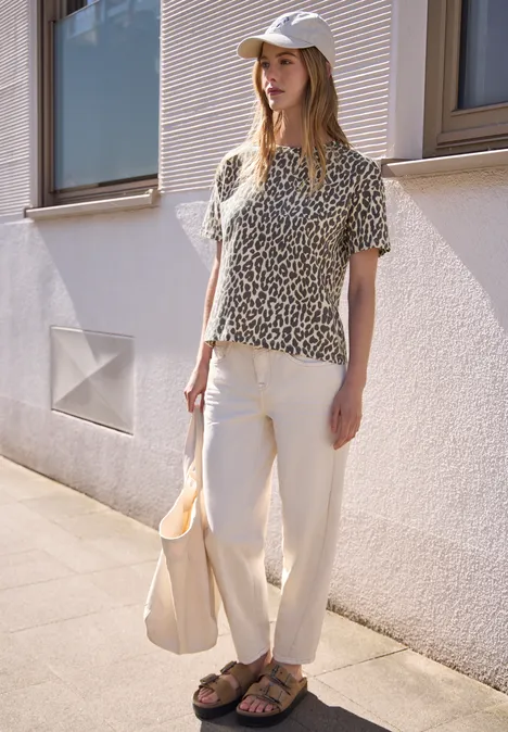 T-Shirt mit Rundhals und Leo-Muster light buffed beige Frau in sommerlichem Outfit mit Leopardenoberteil, weißen Chinos, Sandalen, Beutel und Hut vor Hauswand.