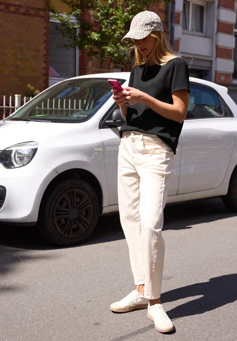 T-Shirt in Unifarbe mit Raffung Black Frau in schwarzem T-Shirt, weißen Hosen und Cap schaut auf ihr Smartphone vor kleinem weißen Auto.