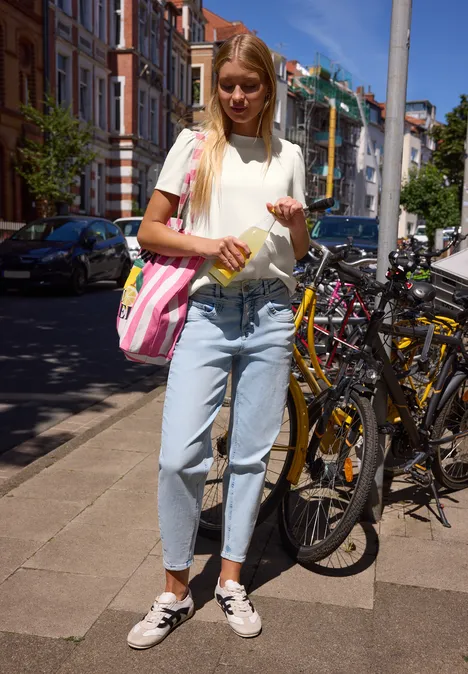 T-Shirt in Unifarbe mit Raffung off white Junge Frau in Weißshirt und Jeans steht auf dem Gehweg neben geparkten Fahrrädern, hält eine Getränkeflasche.