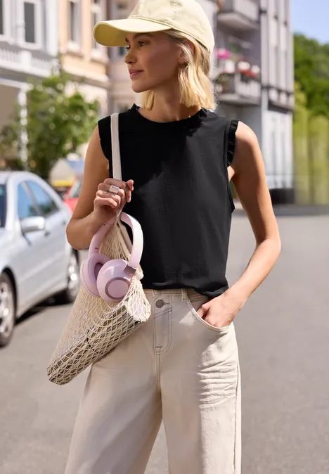 Eine Frau in schwarzem Top, beigen Hosen und Cap geht mit einer geflochtenen Tasche über die Straße.