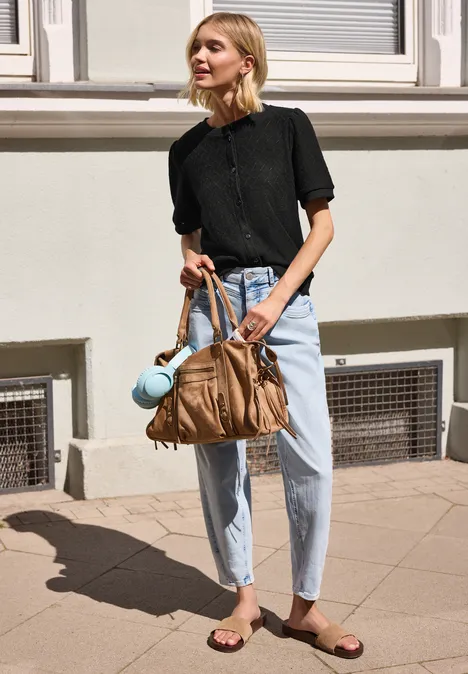 Frau mit schwarzem Oberteil und hellen Jeans steht vor einer Hauswand und hält eine große braune Tasche.