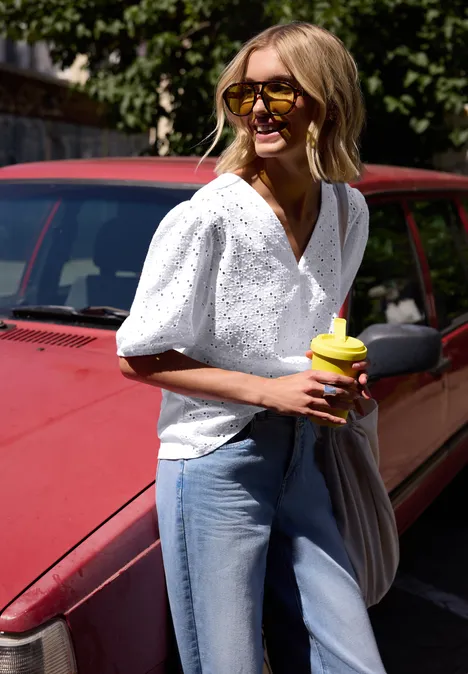 Femme en chemisier blanc et jean debout devant une voiture rouge, tenant un gobelet.