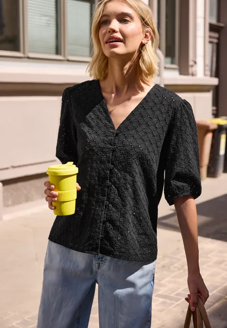 Frau in schwarzer Bluse und Jeans hält gelben Coffee-to-go-Becher auf sonnigem Bürgersteig.