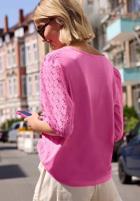 Vrouw in roze top met ajourmouwen staat op straat en bekijkt haar smartphone.