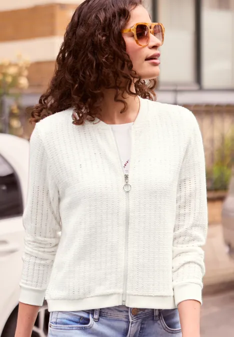 Jas met lange mouwen, structuur en ritssluiting off white Vrouw met krullend haar, zonnebril en lichte cardigan, die opzij kijkt in een stedelijke omgeving.
