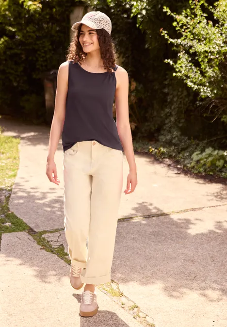 Asymmetrische top met ronde hals very berry Jonge vrouw loopt op een zonnig pad, draagt zwart hemdje, lichte broek, witte sneakers en een pet.