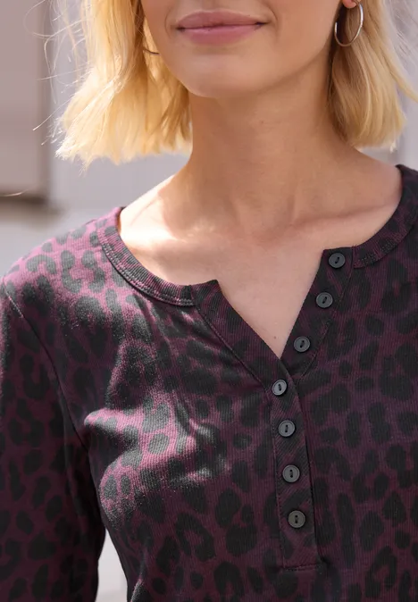 Nahaufnahme einer Frau mit kurzem blonden Haar, die ein lila Shirt mit Leopardenmuster und Knopfleiste trägt.