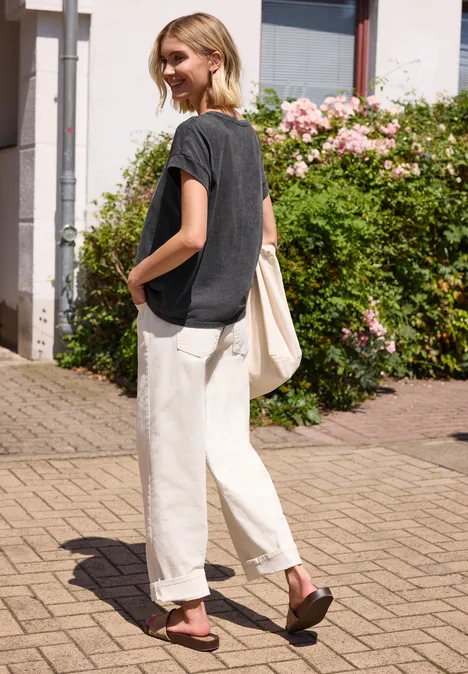 Eine Frau im schwarzen T-Shirt und beigen Hose mit Tragetasche steht auf Kopfsteinpflaster vor blühendem Strauch.