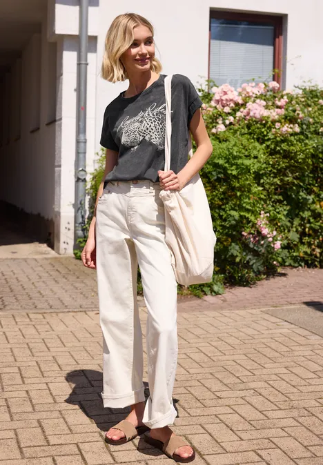 Person in T-Shirt, hellen Hosen und Sandalen auf dem Bürgersteig vor einem Haus mit Rosen trägt weiße Tasche.