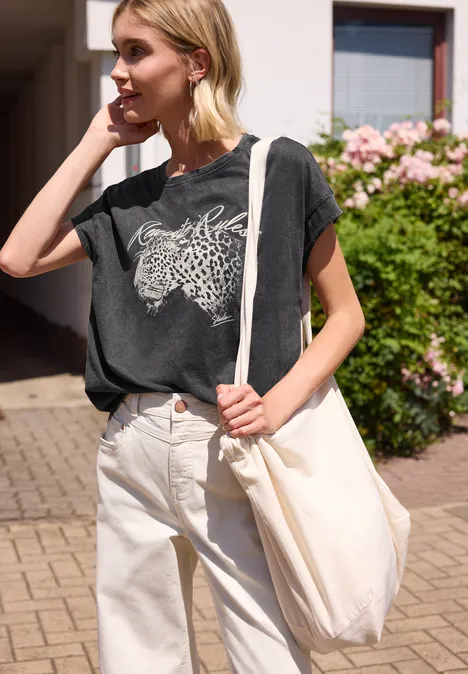 Eine Person trägt draußen ein graues T-Shirt mit Tiermotiv, helle Hose und eine weiße Stofftasche.