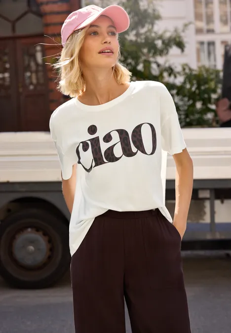 Eine Person steht auf der Straße vor einem Bus, trägt weißes T-Shirt mit 'ciao' und schwarze Hose.