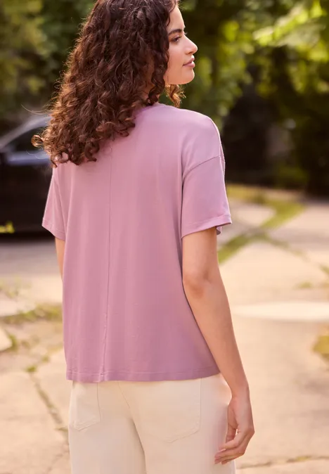 T-shirt à col rond avec inscription pearly rose Femme vue de dos, cheveux bouclés, t-shirt rose et pantalon clair, debout sur un chemin au milieu de verdure.
