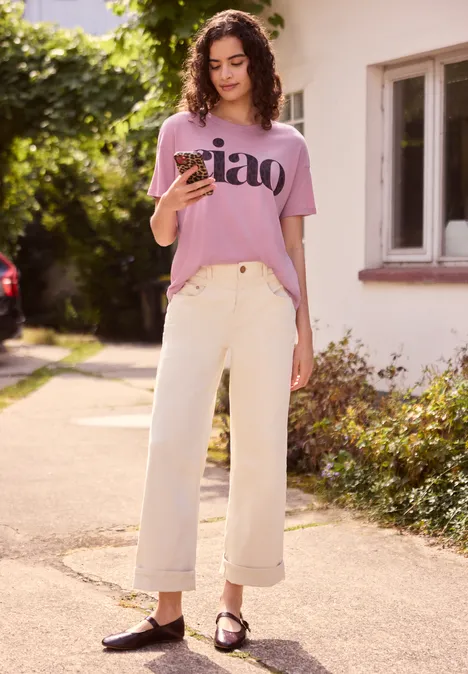 T-shirt à col rond avec inscription pearly rose Femme debout devant une maison, regarde son smartphone, porte un t-shirt rose et un pantalon blanc.