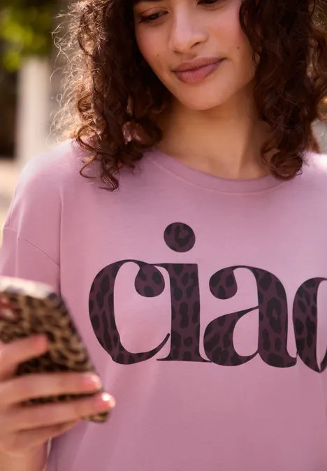 T-shirt à col rond avec inscription pearly rose Femme en t-shirt rose 'ciao', souriante, regardant son smartphone à coque léopard.
