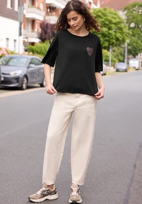 Person mit lockigen Haaren steht in ruhiger Wohnstraße, trägt schwarzes Shirt mit Herz, helle Hose und Sneakers.