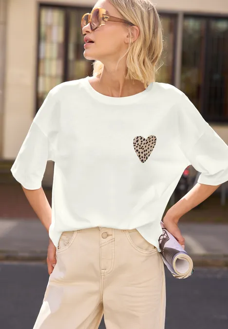 Frau in weißem T-Shirt mit kleinem Herz-Leo-Print, beiger Hose, hält eine zusammengerollte Zeitschrift.