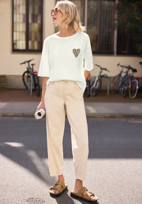 Eine Frau steht auf der Straße, trägt ein weißes Oberteil mit Herzmotiv, beige Hose und braune Sandalen.