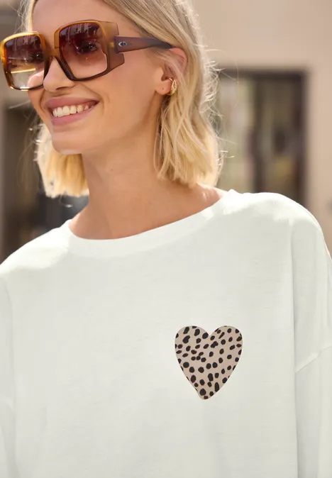 Frau trägt weißes T-Shirt mit kleinem, gepunktetem Herzmotiv auf der Brust.