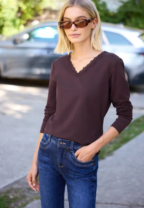 Vrouw met zonnebril, donkerbruine longsleeve en blauwe jeans, staand op de stoep, hand in zak.
