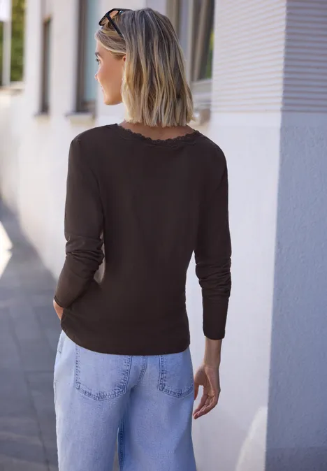 Vrouw van achter in zwart shirt en lichtblauwe jeans, blond haar en zonnebril op het hoofd, bij een witte muur.